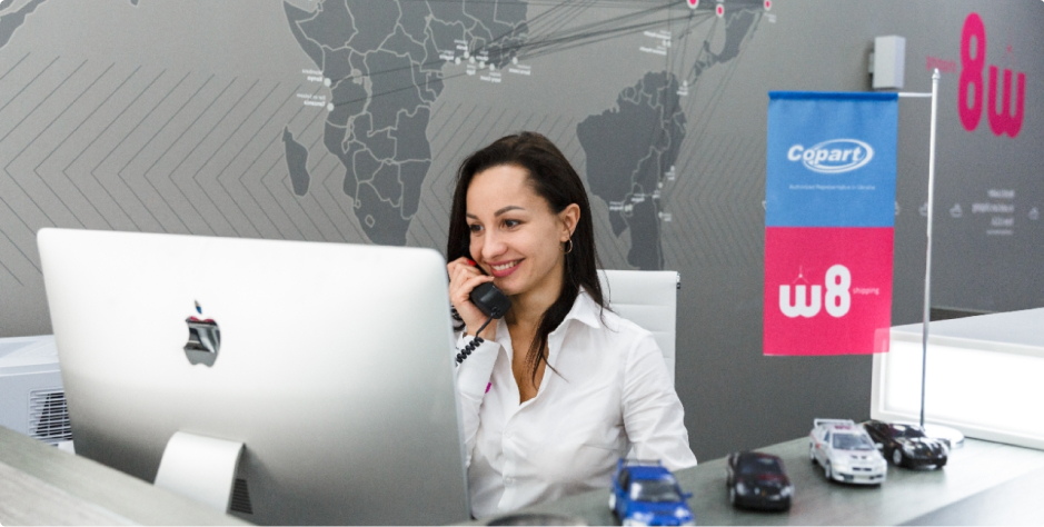 Uśmiechnięta kobieta w białej koszuli rozmawia przez telefon, siedząc przy biurku z laptopem Apple w biurze z mapą świata na ścianie i flagą z logo "Copart" oraz "W8 shipping" w tle. Na biurku przed nią znajdują się modele samochodów.